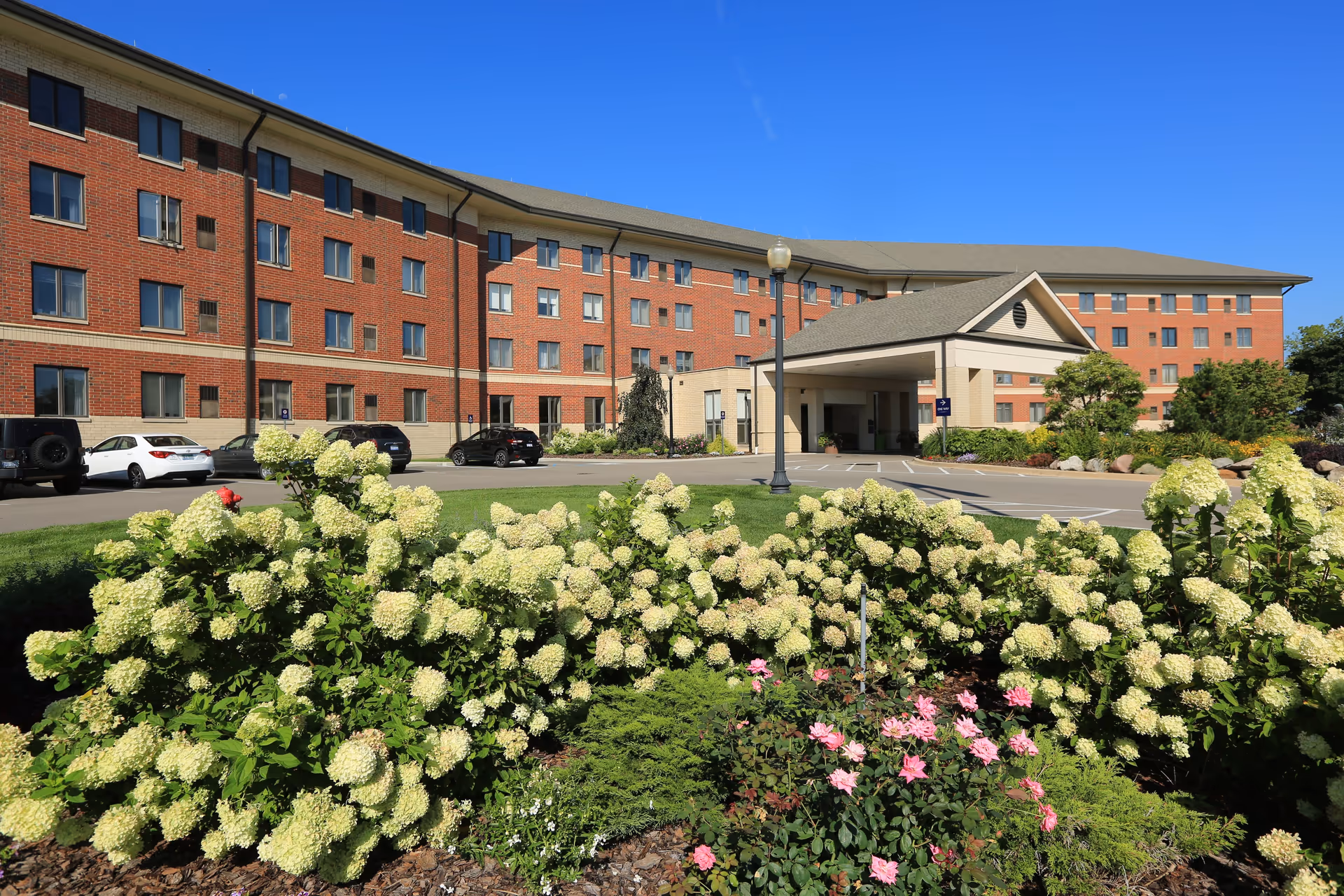 Exterior view of a large brick retirement community building with multiple windows, a covered entrance, a parking lot with several cars, and landscaped flower beds with white and pink flowers in the foreground under a clear blue sky.