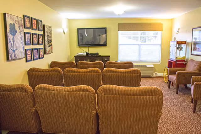 A cozy senior living facility common room with several brown upholstered chairs arranged in rows facing a wall-mounted flat screen TV. The room has beige walls decorated with framed pictures and a popcorn machine near a window with blinds. The floor is carpeted in a patterned design.