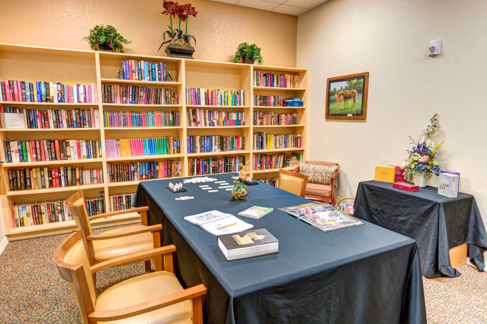 A cozy room with a large bookshelf filled with books along the back wall. In front of the bookshelf is a rectangular table covered with a black tablecloth, holding books, magazines, and a small plant. There are wooden chairs with cushioned seats around the table. To the right, there is a smaller table also covered with a black tablecloth, decorated with a flower arrangement and some boxes. A framed painting of a horse hangs on the wall above a cushioned chair with a blanket draped over it.