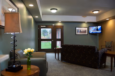 A cozy living room area with a green armchair and a side table holding a lamp and a vase of yellow flowers. Across from the armchair is a dark patterned sofa facing a wall-mounted television showing a news channel. There is a small wooden table between the sofa and the glass exit doors, which provide a view of green grass and trees outside. The walls are painted green and beige, and there is a framed picture hanging on the wall next to the TV.