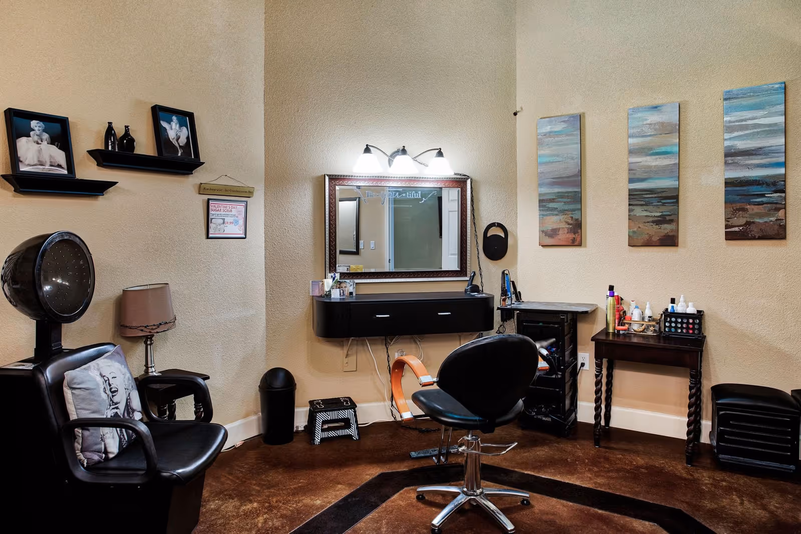 Interior of a salon room with a black salon chair in front of a wall-mounted mirror and vanity. To the left is a black hair dryer chair with a Marilyn Monroe pillow, a small table with a lamp, and two framed photos on the wall. On the right side, there is a small table with hair products and three abstract paintings hanging on the wall.