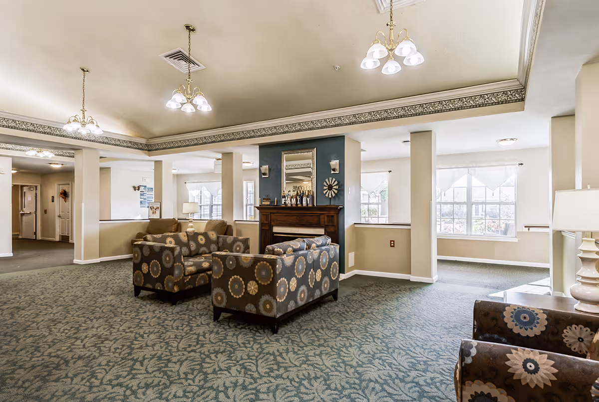 A spacious and well-lit common area in a senior living facility featuring patterned armchairs arranged around a wooden fireplace with a mirror and decorative items on the mantel. The room has large windows with white curtains, green patterned carpet, beige walls, and ceiling lights with multiple bulbs.