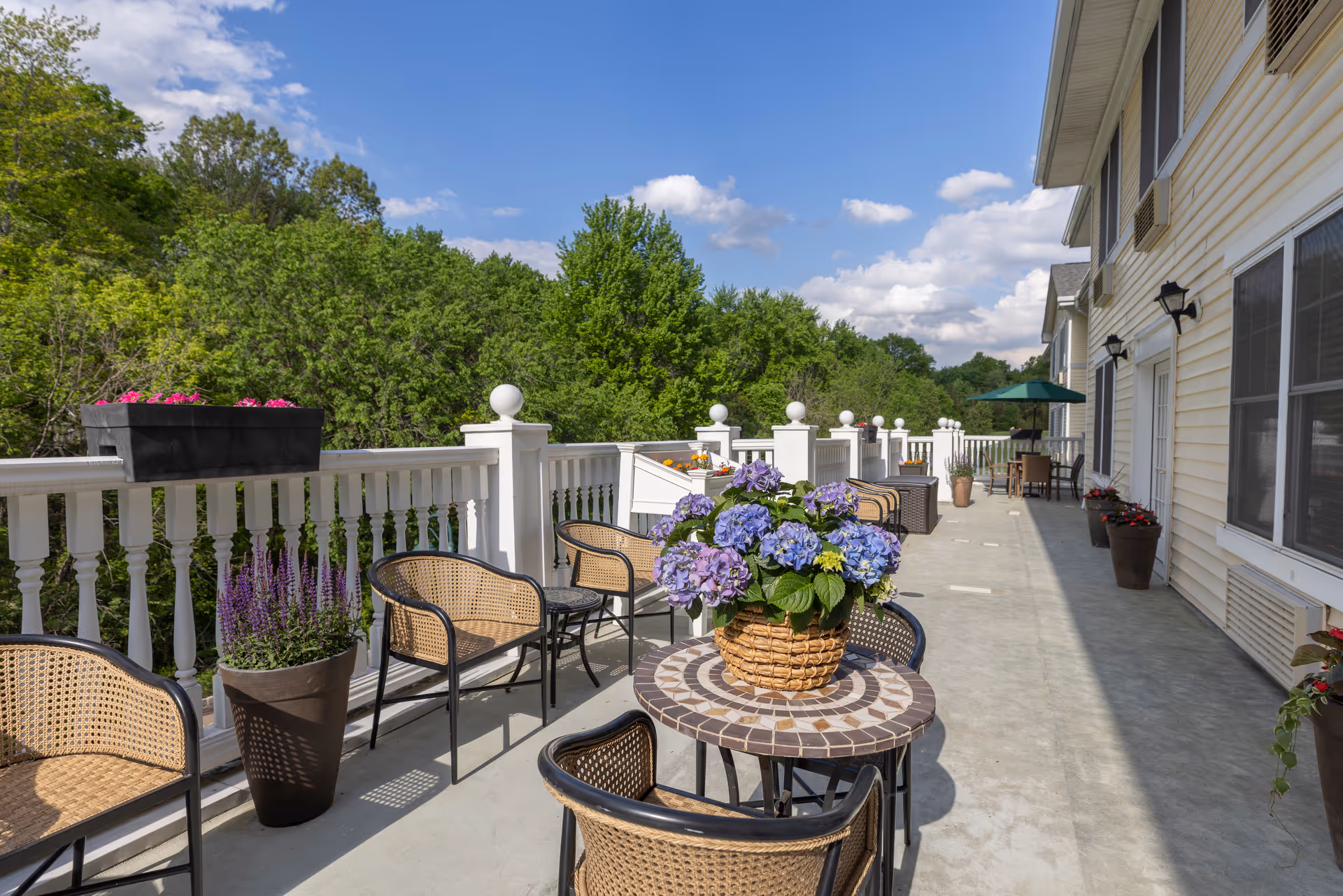 Outdoor patio area at Mira Vie at Green Knoll Assisted Living Bridgewater featuring a tiled round table with a basket of purple and blue flowers, surrounded by wicker chairs. The patio has white railing with decorative posts and several potted plants along the edge. The building exterior is light-colored with windows and outdoor wall lights. Trees and a blue sky with clouds are visible in the background.