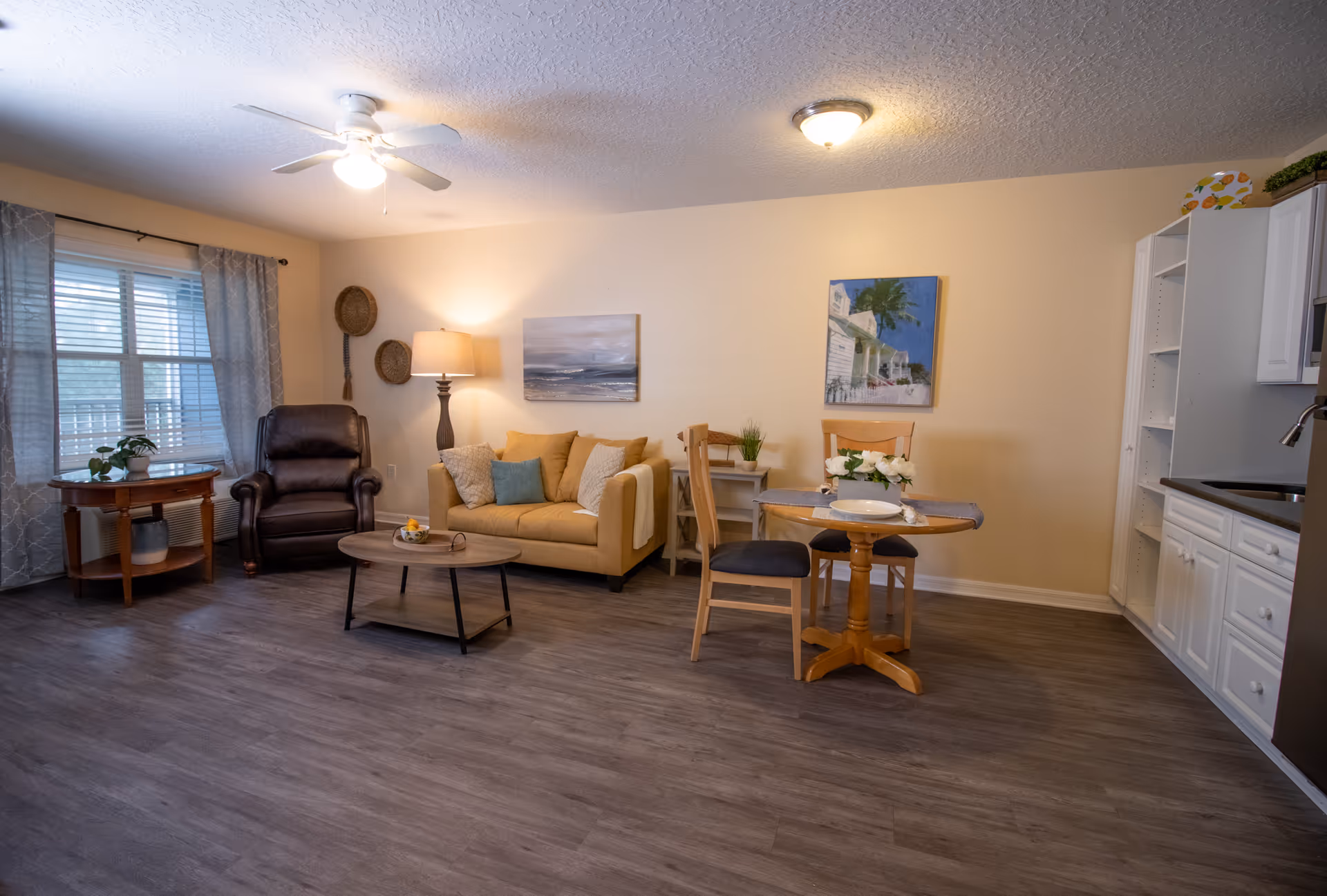A cozy living room and dining area with a beige couch, a brown leather armchair, a wooden coffee table, and a small round dining table with two chairs. The room has wood flooring, light yellow walls, two paintings on the wall, a window with patterned curtains, a floor lamp, and a ceiling fan with a light. The kitchen area with white cabinets and a sink is partially visible on the right.
