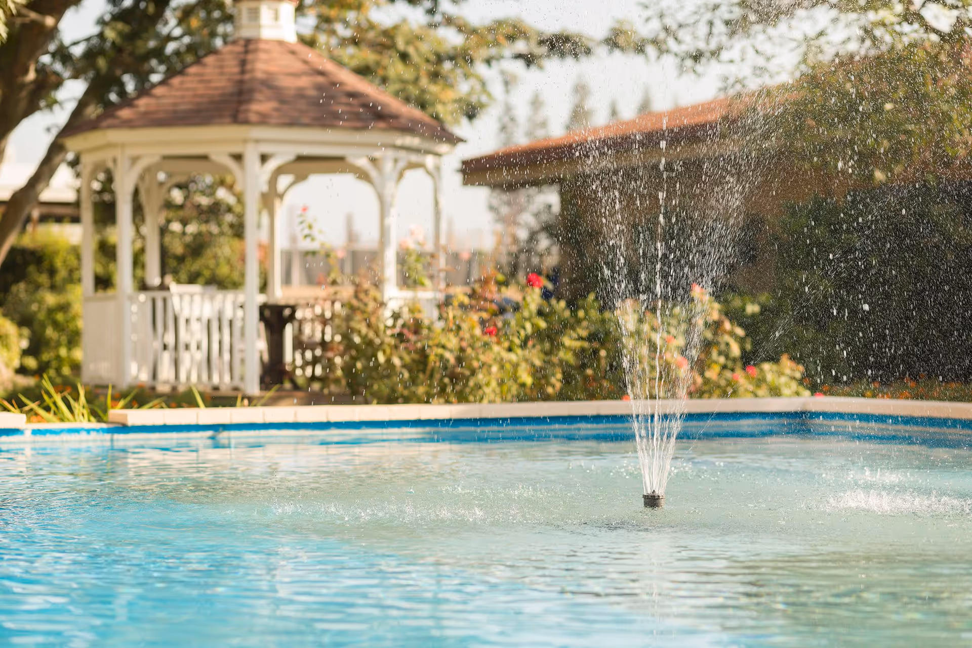 A water fountain spraying water in a pool with a white gazebo and greenery in the background on a sunny day.