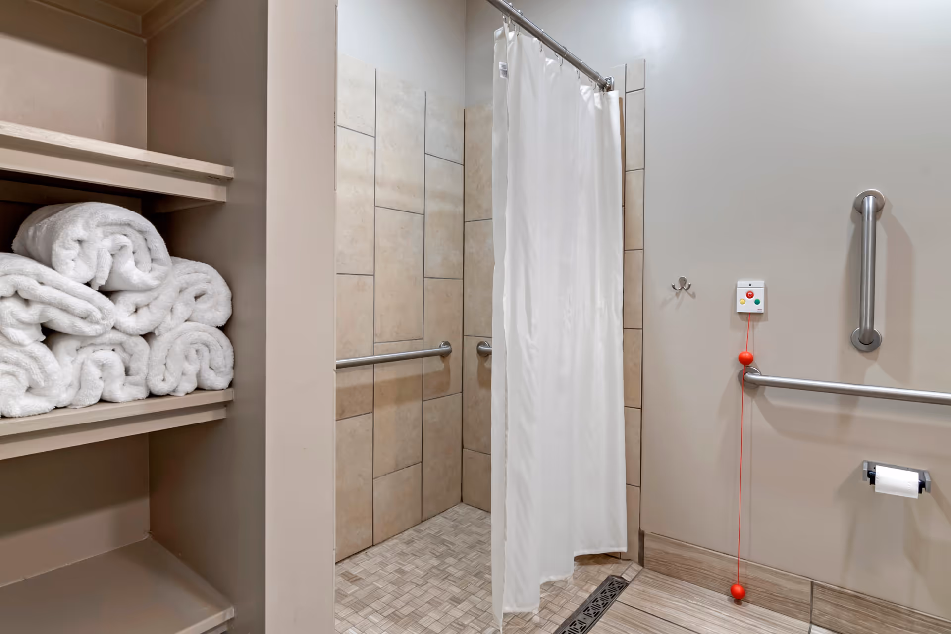 Accessible bathroom with a tiled walk-in shower behind a white curtain, wall grab bars, towel shelves stacked with white towels, and an emergency pull cord.