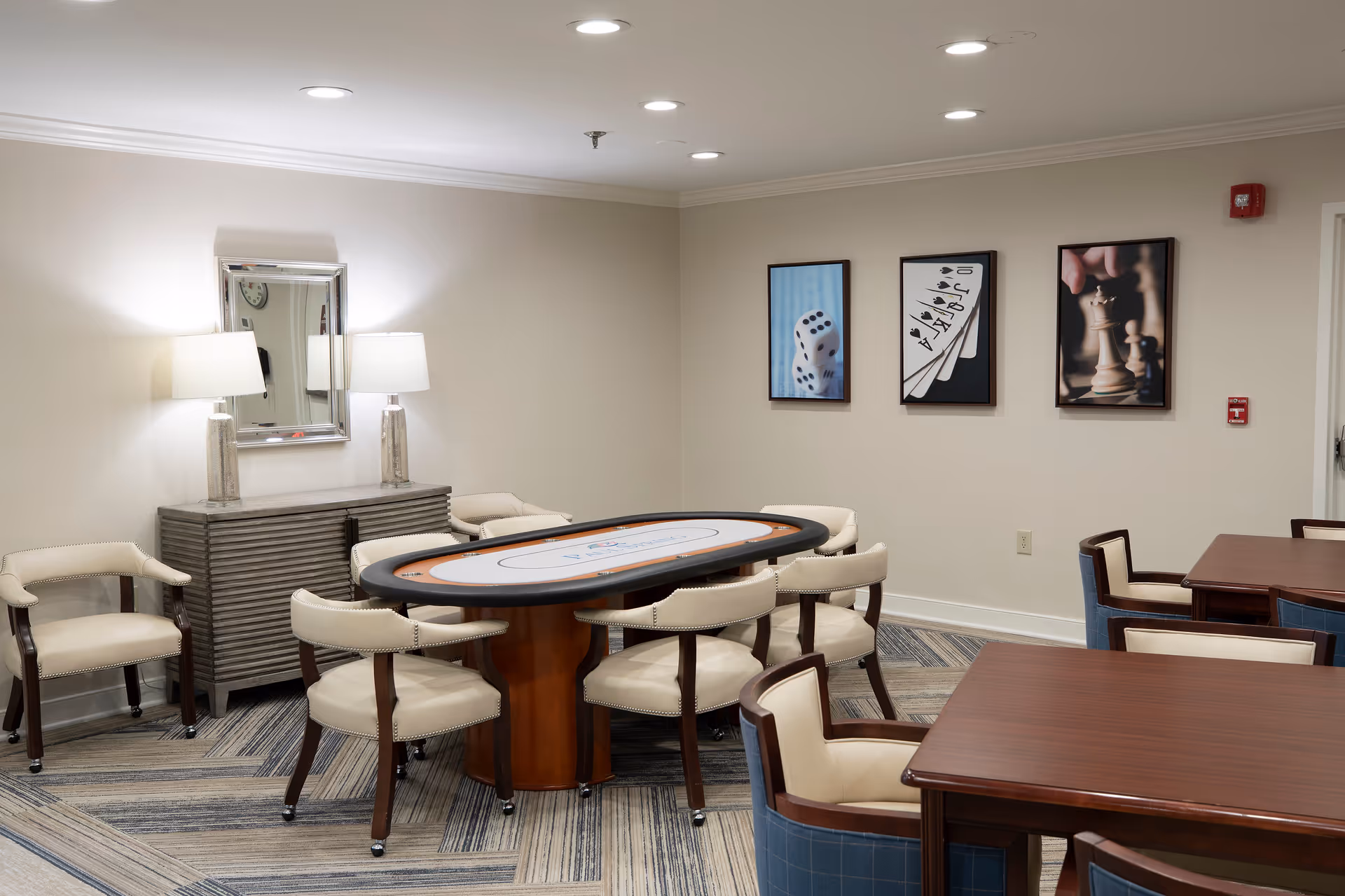A well-lit room with a poker-style gaming table surrounded by cream-colored chairs with wooden legs. There are additional wooden tables and chairs in the foreground. On the back wall, three framed pictures depict dice, playing cards, and chess pieces. A sideboard with two lamps and a mirror is positioned against the left wall. The room has a patterned carpet and recessed ceiling lights.