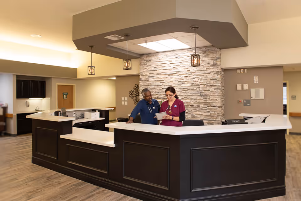 Two healthcare workers standing behind a large reception desk in a well-lit facility with a stone accent wall and pendant lights overhead. One worker is wearing blue scrubs and the other is wearing maroon scrubs, and they are looking at a tablet together.