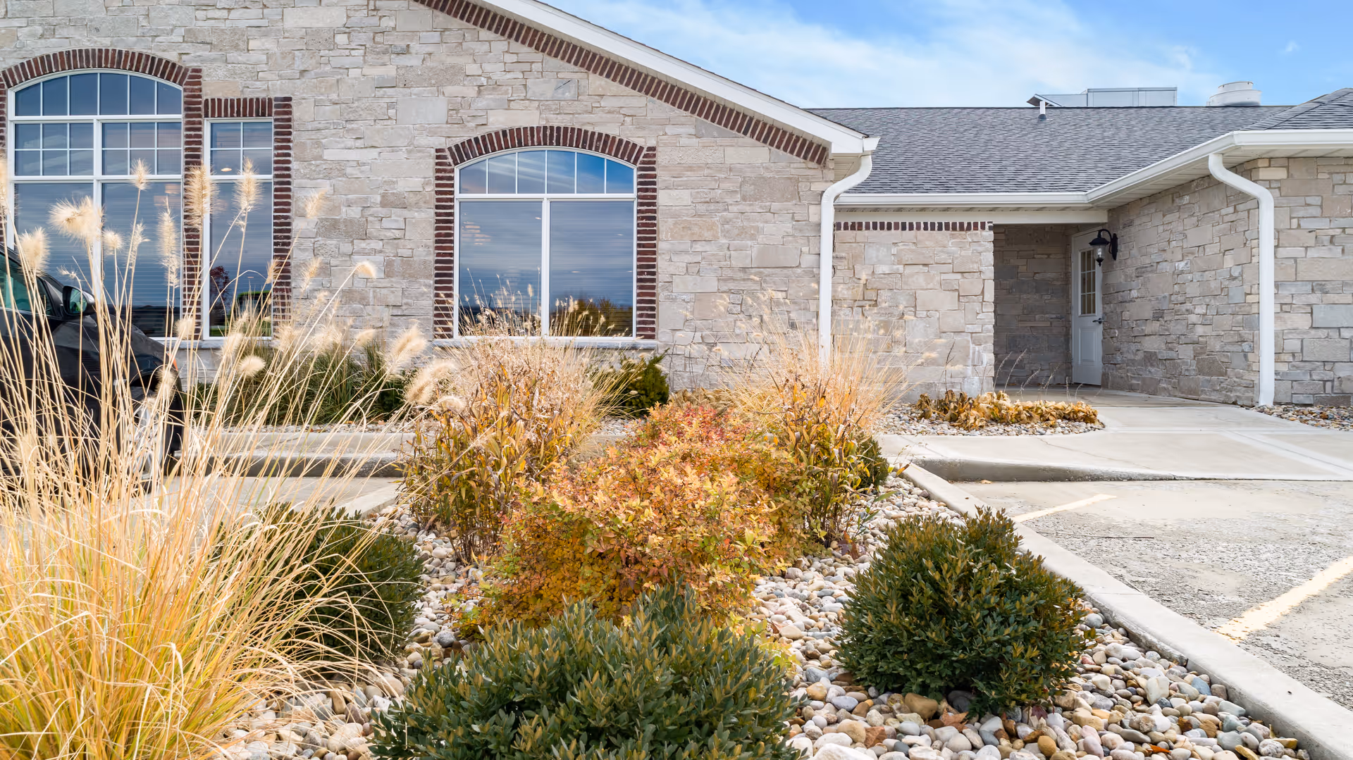 Exterior view of a stone building with large windows and a landscaped area featuring ornamental grasses and shrubs in front. The sky is clear with some clouds.