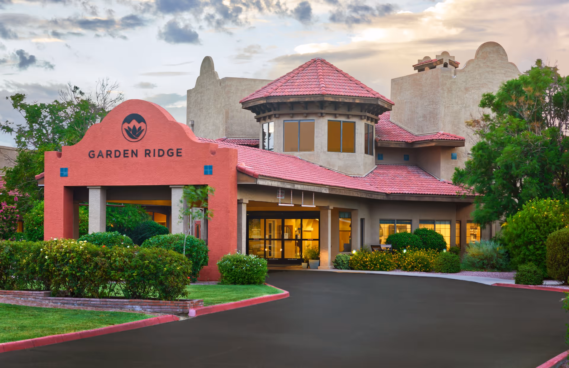 Exterior view of the Garden Ridge senior living facility building with a red tiled roof, beige walls, and a prominent red entrance archway displaying the Garden Ridge name and logo. The entrance is surrounded by well-maintained green bushes and trees, with a paved driveway leading up to the building under a partly cloudy sky.