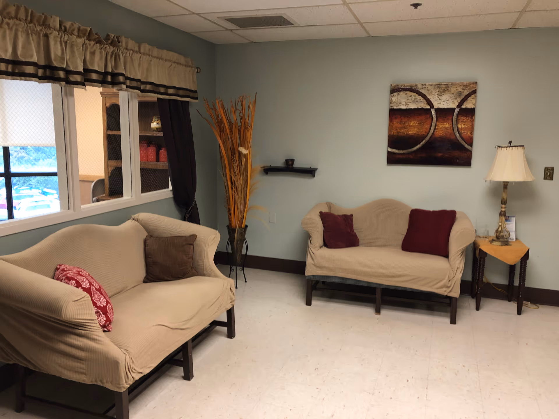 A small sitting area with two beige loveseats covered with slipcovers, each adorned with decorative pillows. There is a small wooden side table with a lamp and a yellow cloth on it. A piece of abstract wall art featuring circular designs hangs on the light blue wall. A tall vase with decorative dried plants stands in the corner near a window with beige and brown valance curtains.