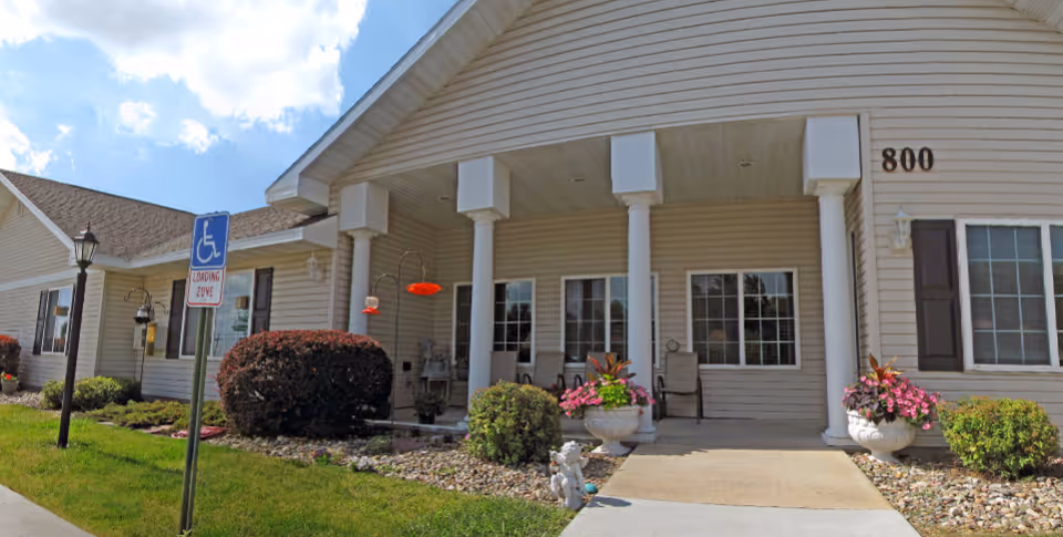 Exterior view of a beige senior living facility building with white columns at the entrance, a wheelchair accessible loading zone sign, well-maintained bushes, flower pots with pink flowers, and a clear blue sky with some clouds.