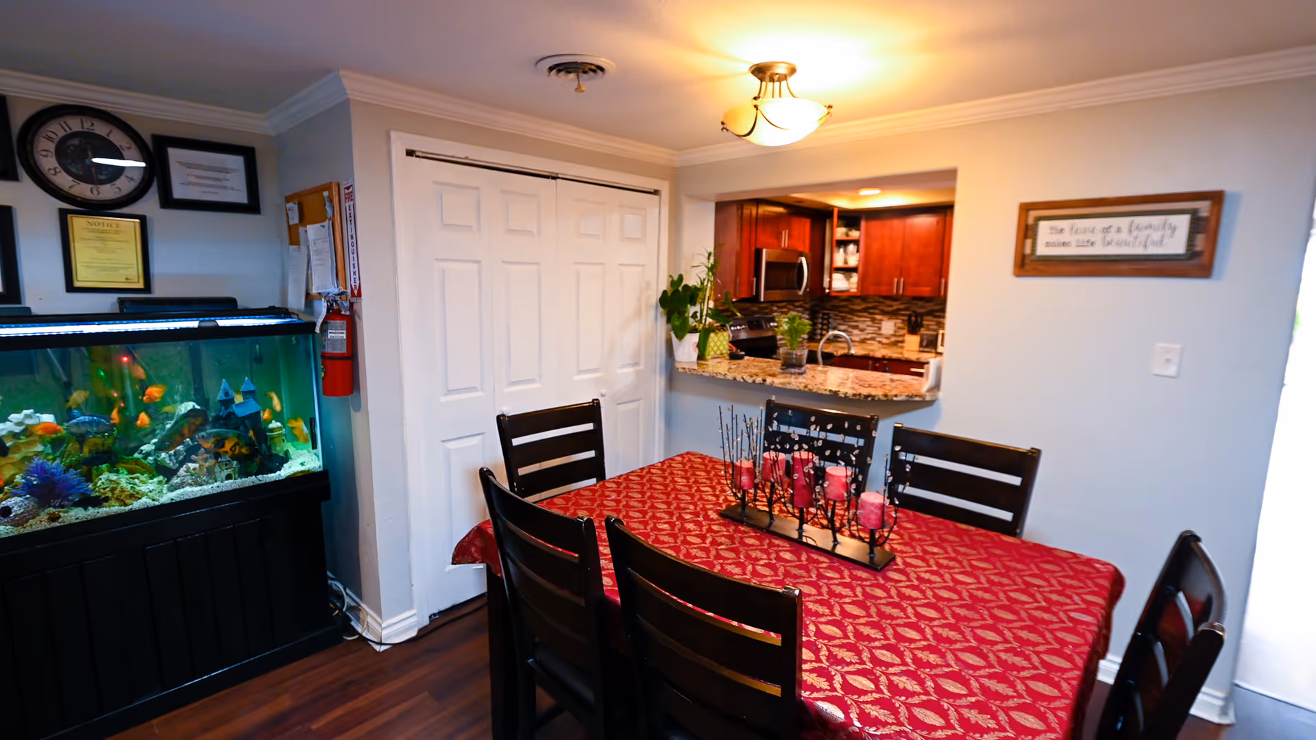 Dining room with a red patterned tablecloth on a table surrounded by dark chairs, a large aquarium against the left wall, and a kitchen visible through a pass-through.