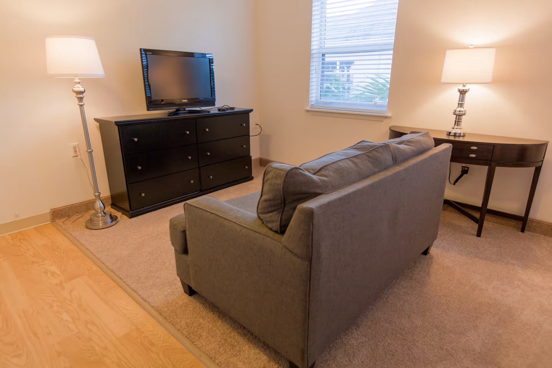 A small living room area with a gray couch facing a black dresser with a flat-screen TV on top. There is a floor lamp to the left of the dresser and a table lamp on a dark wooden console table to the right near a window with blinds.