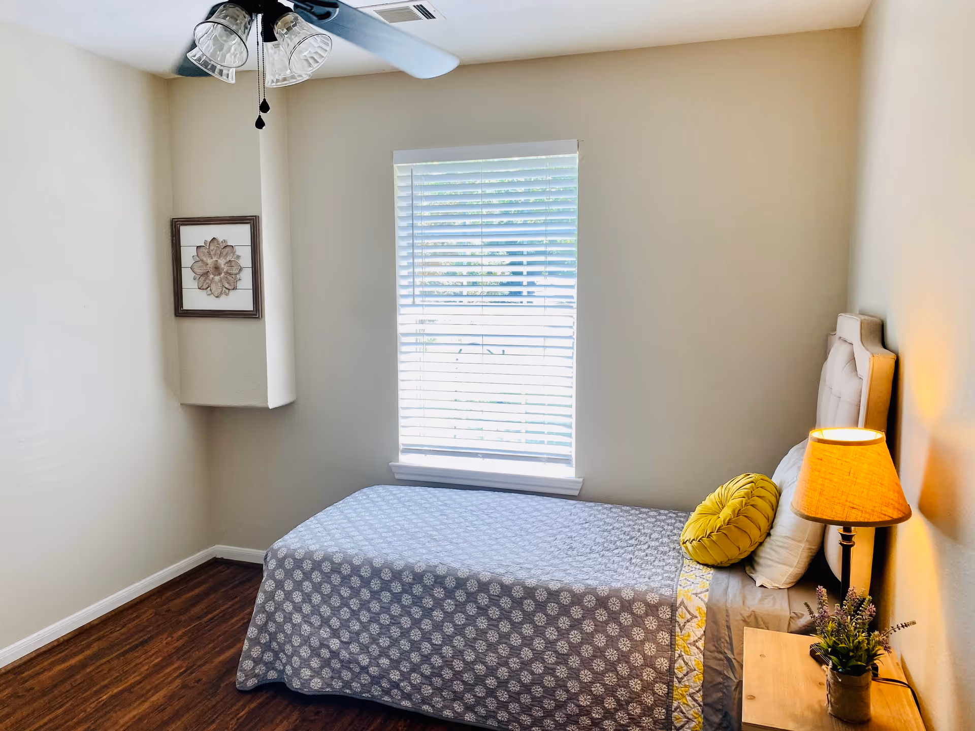 A small bedroom with a single bed covered in a gray patterned bedspread, a yellow round pillow, and a white pillow. There is a wooden nightstand with a lamp and a small potted plant next to the bed. A window with white blinds is centered on the wall opposite the bed. A ceiling fan with lights is mounted on the ceiling, and a framed flower artwork hangs on the left wall. The room has wooden flooring and light-colored walls.