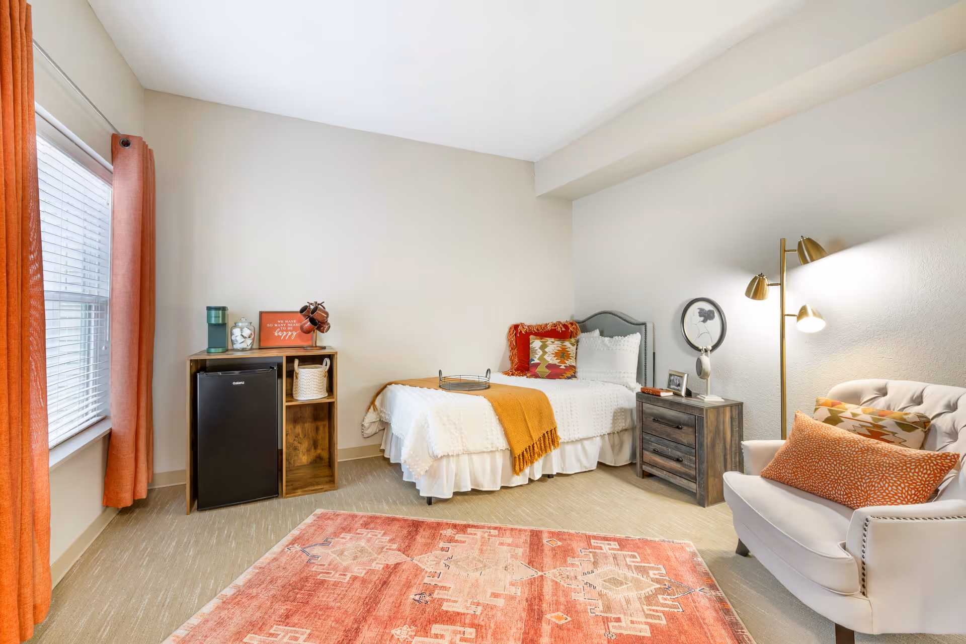 A cozy bedroom in an assisted living facility featuring a single bed with white bedding and colorful pillows, a wooden nightstand with a lamp and decorative items, a comfortable armchair with patterned cushions, a small wooden cabinet with a mini fridge, and a window with orange curtains. The room has light-colored walls and flooring, and a patterned area rug in warm tones.