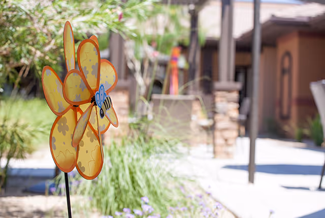 Yellow flower-shaped pinwheel garden decoration with a bee in a courtyard with plants and a building in the background.