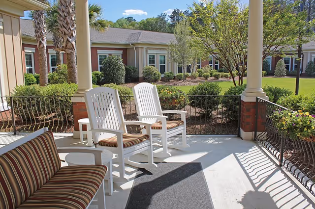 Covered outdoor patio with white rocking chairs, striped cushioned seating, and a view of a landscaped courtyard and building.