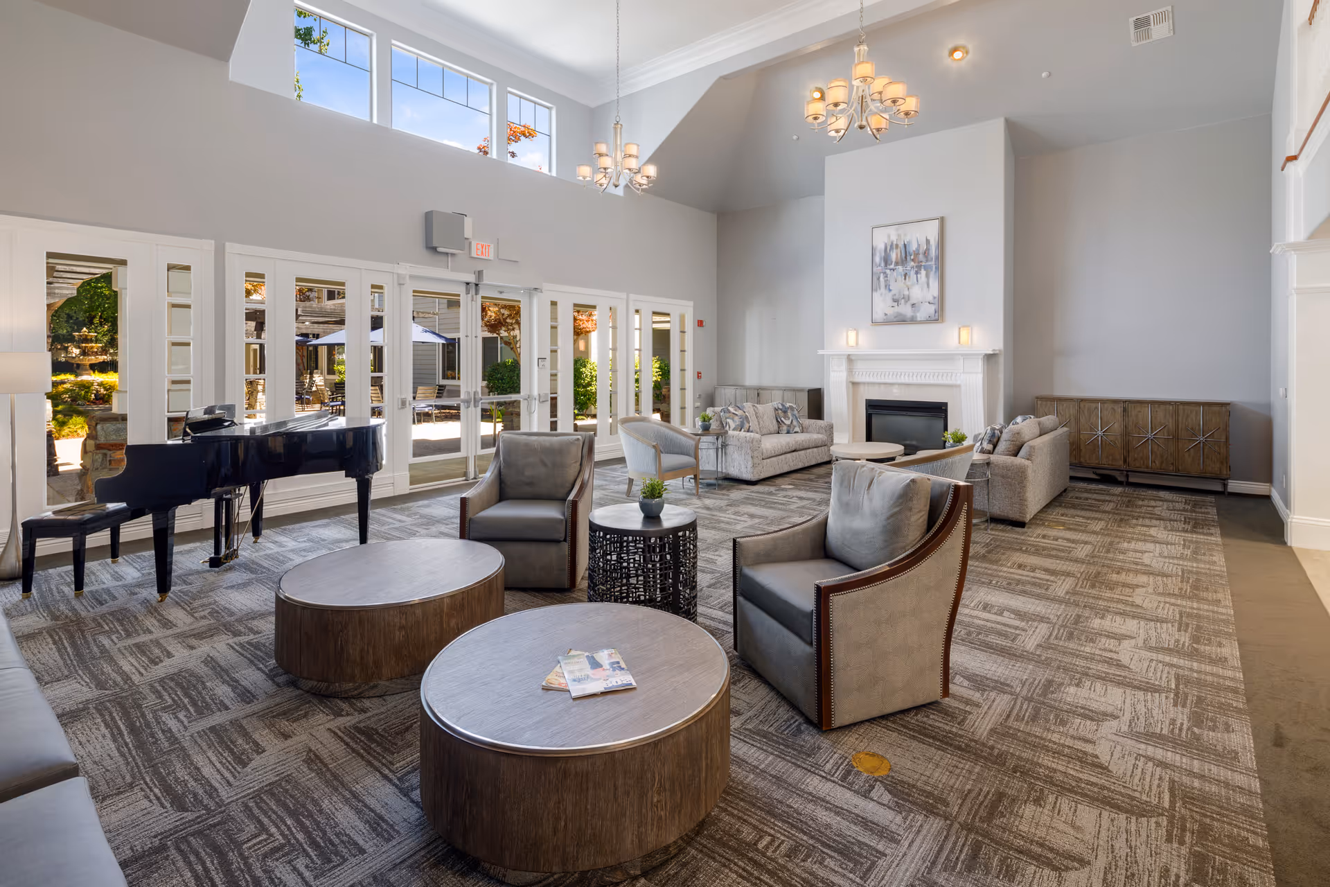 Bright, spacious communal lounge with sofas, armchairs, round coffee tables, a grand piano, and a fireplace under tall windows and chandeliers.