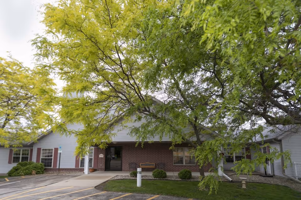 Exterior view of McFarland Villa Assisted Living facility showing a single-story building with white siding and brick facade, surrounded by green trees and a parking area in front.