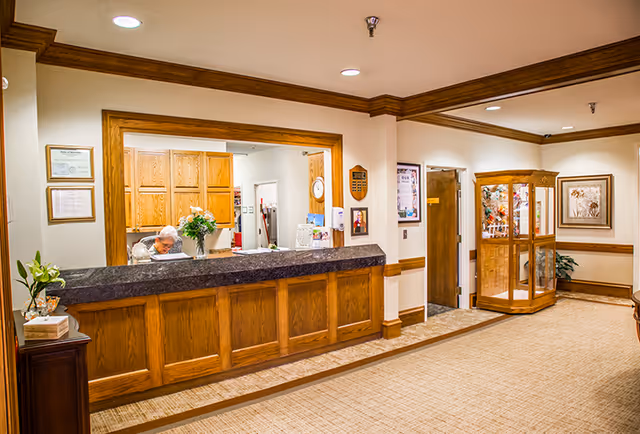 Reception area of Westgate Assisted Living with a wooden front desk, a person sitting behind the desk, wooden cabinetry in the background, framed certificates on the wall, a glass display cabinet, and beige carpeted flooring.