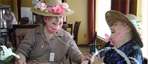 Two elderly women wearing decorative straw hats with flowers are sitting at a table in a well-lit room, smiling and holding hands. One woman is wearing a beige top and the other a dark floral top. There are chairs and other people visible in the background.