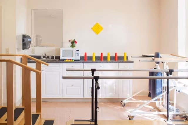 A rehabilitation or therapy room with parallel bars for walking practice, a small kitchenette area with a microwave, sink, and countertop, and a wooden staircase with handrails. The room is well-lit with natural light and has a clean, minimalistic design.