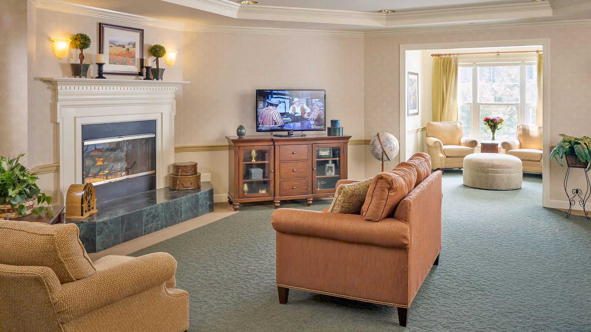 A cozy living room with a brown sofa facing a wooden TV stand with a flat-screen television. To the left is a fireplace with a white mantle decorated with two small plants and a framed picture. There is a beige armchair in the foreground and a green carpet covering the floor. In the background, a sunlit seating area with two beige armchairs, a round ottoman, and a small table with a vase of flowers is visible near large windows with yellow curtains.