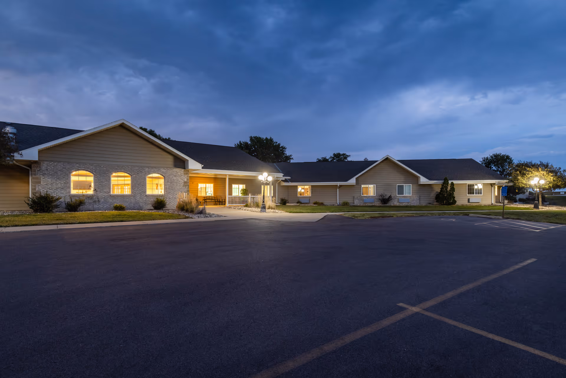 Exterior view of a single-story senior living facility building at dusk with lights glowing from the windows and a mostly empty parking lot in front.
