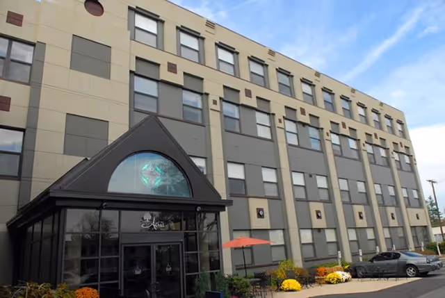 Front entrance of a multi-story senior living building with a glass canopy, rows of windows, a small patio with an umbrella and a parked car.