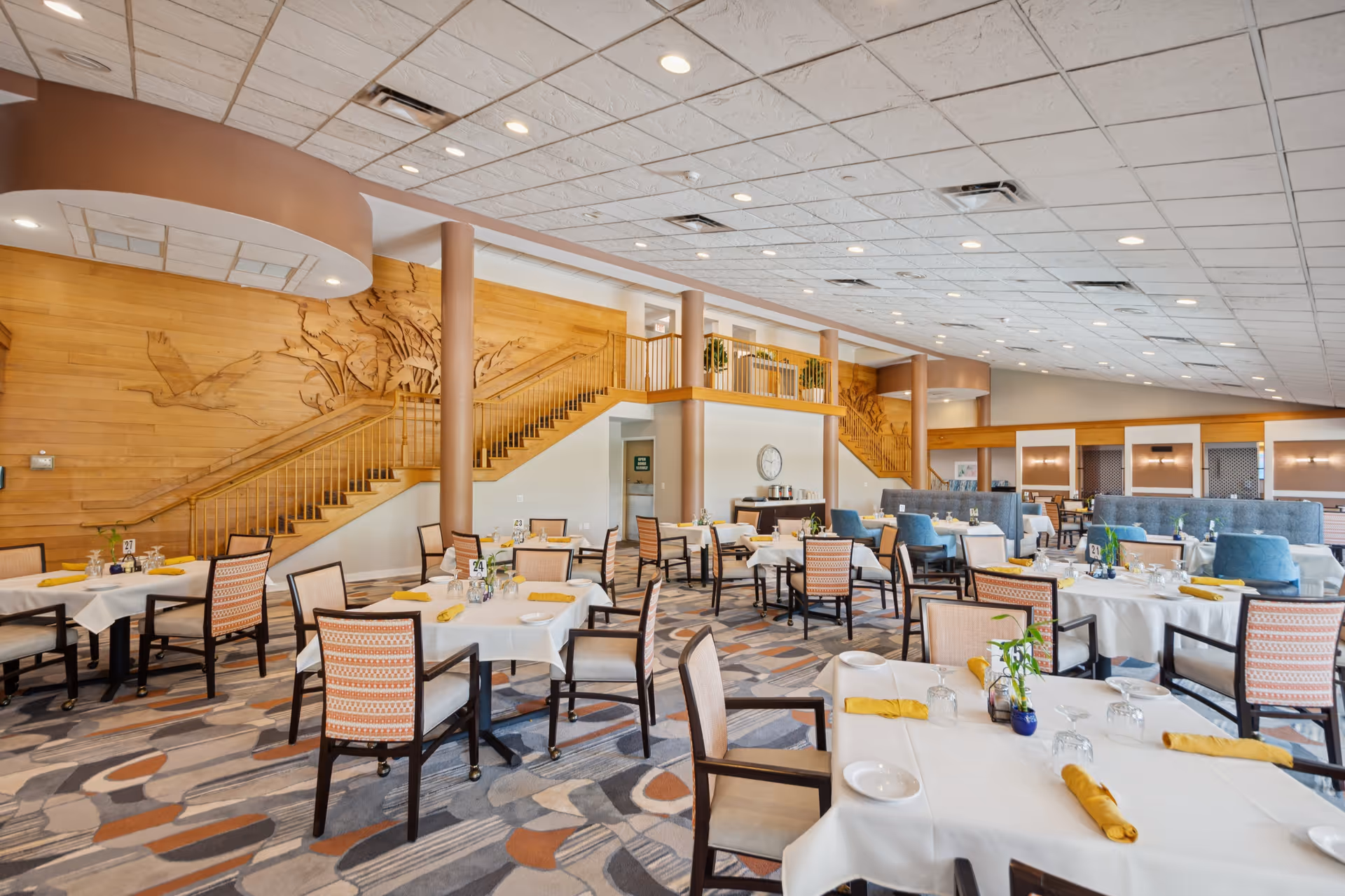 Spacious dining room with multiple tables covered in white tablecloths, each set with plates, glasses, and yellow napkins. The room features patterned carpet flooring, wooden wall paneling with artistic carvings, and a staircase leading to an upper level. Comfortable chairs with patterned upholstery and blue cushioned seating are arranged around the tables. The ceiling has recessed lighting and air vents.