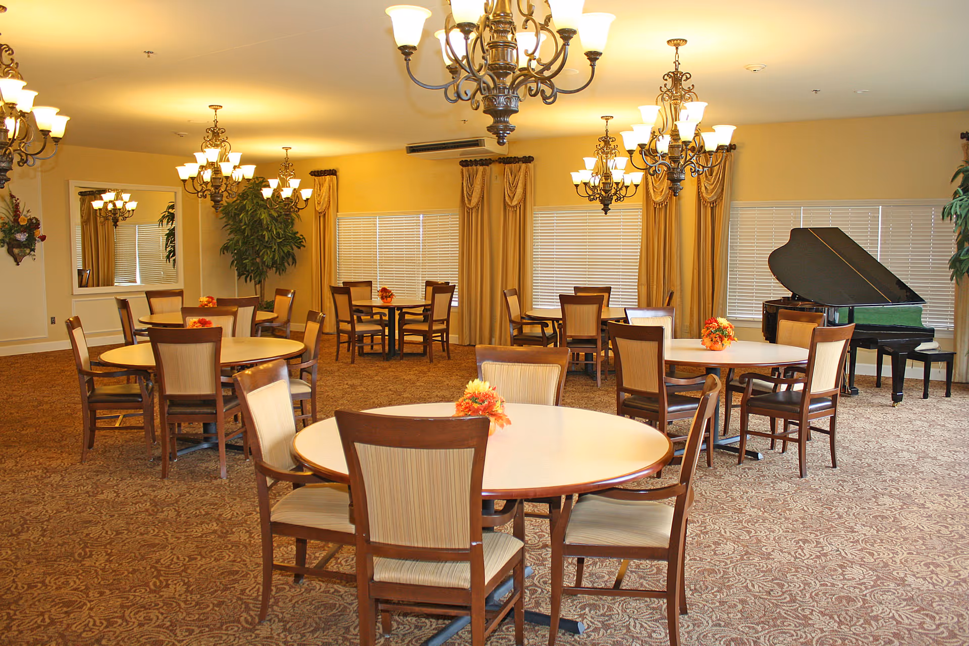 A spacious dining room with multiple round tables and wooden chairs arranged neatly. Each table has a small floral centerpiece. The room is warmly lit by several ornate chandeliers hanging from the ceiling. Large windows with beige curtains line the back wall, and a black grand piano is positioned in the corner. The carpet has a detailed pattern, and there are a few large potted plants adding greenery to the space.