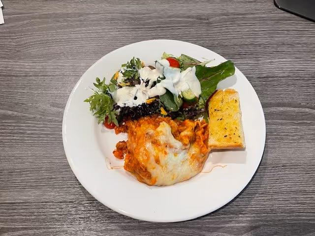 A white plate with a serving of baked pasta topped with melted cheese, a side salad with leafy greens and cherry tomatoes drizzled with dressing, and a piece of garlic bread on a gray wooden table.