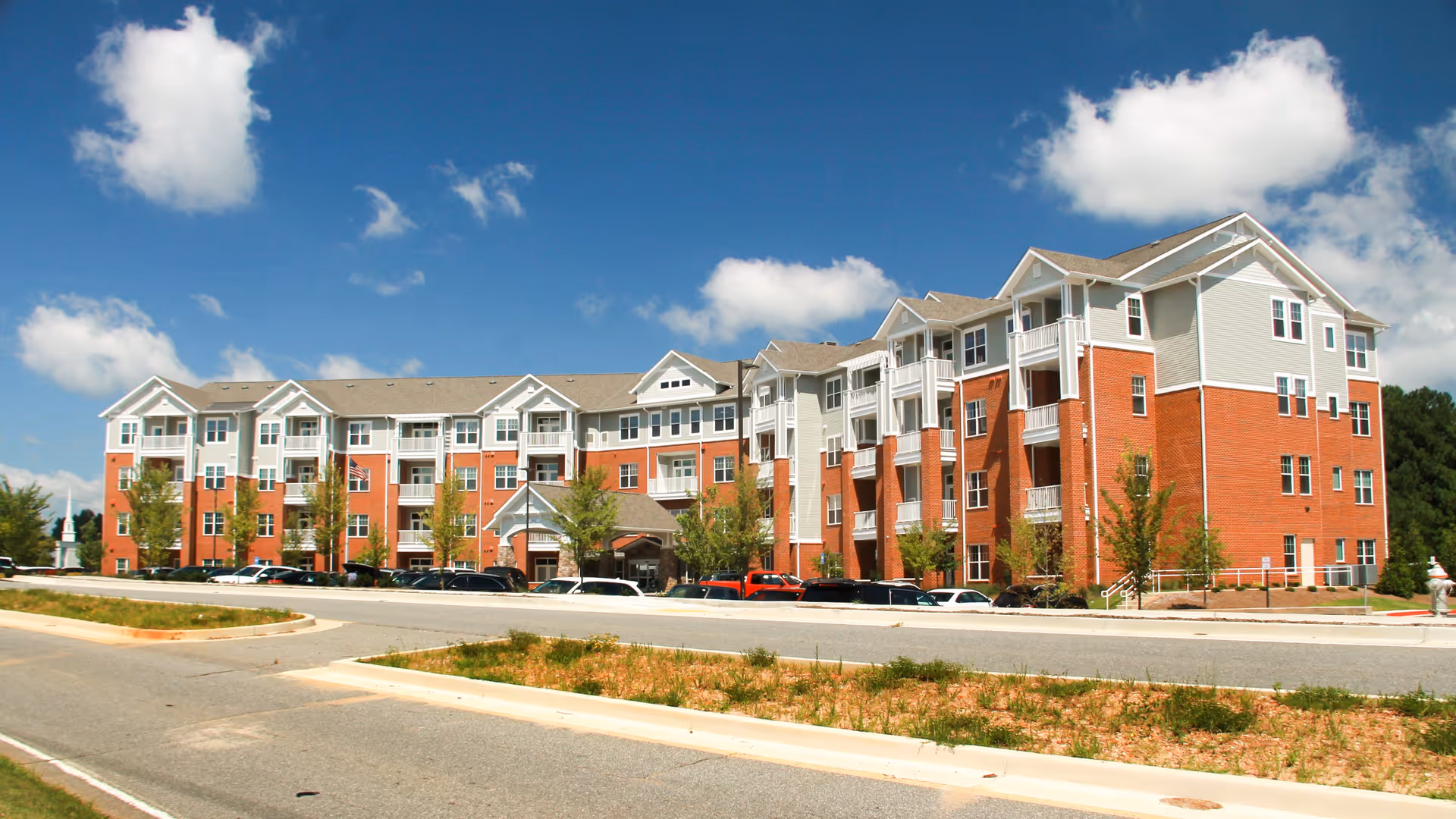 Exterior view of a large, multi-story senior living facility building with red brick and light gray siding under a blue sky with scattered clouds. Several cars are parked in front of the building along a paved road.