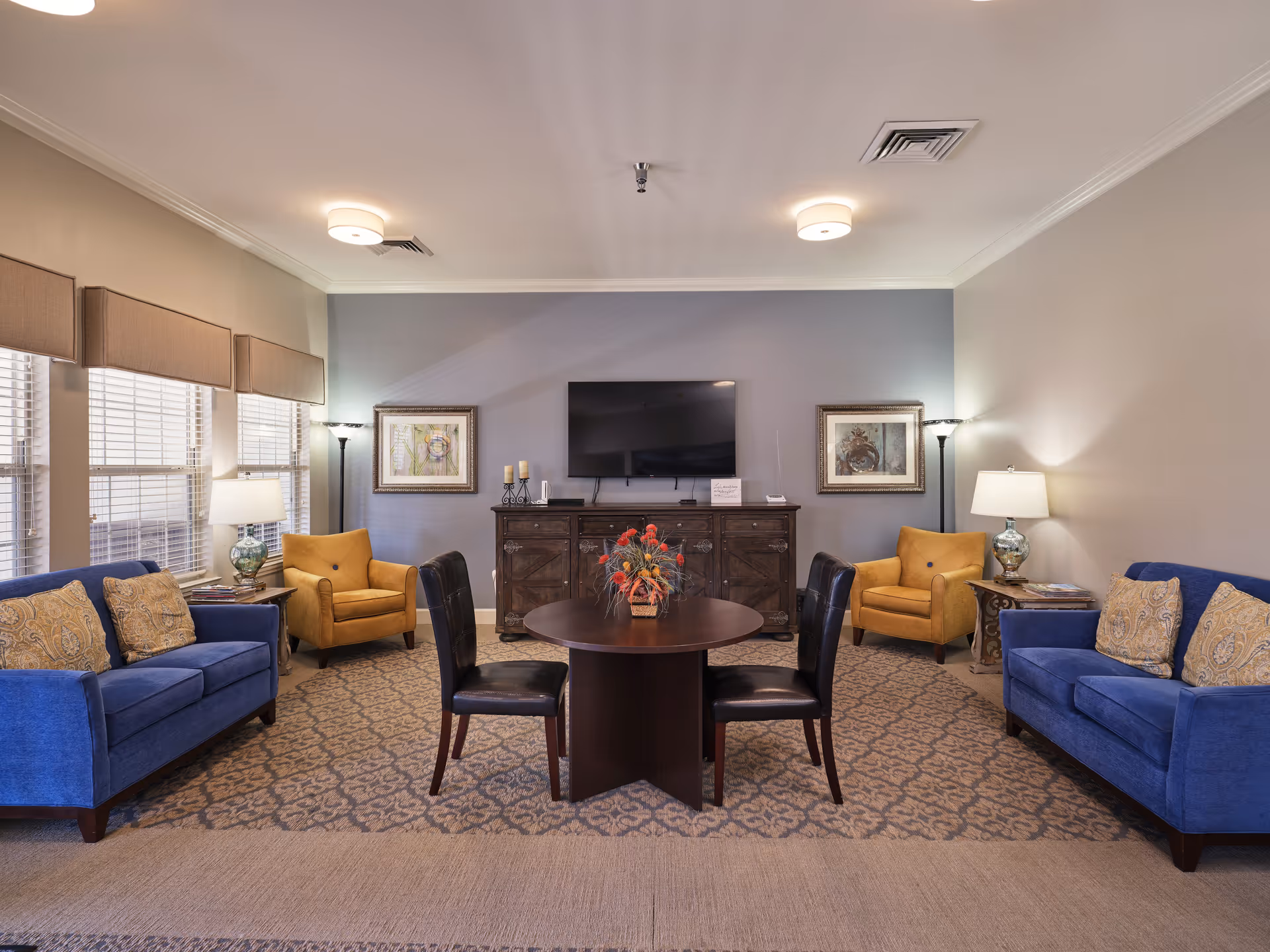 A cozy living room with two blue sofas and two mustard yellow armchairs arranged around a round wooden table with a floral centerpiece. The room features a patterned carpet, two floor lamps, two table lamps on side tables, framed artwork on the walls, and a flat-screen TV mounted above a wooden cabinet.