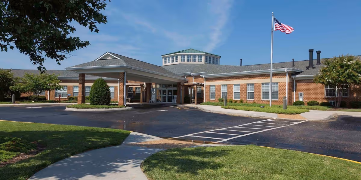 Exterior front view of a single-story brick building with a covered entrance, an American flag on a flagpole, and a paved driveway with parking spaces. The building is surrounded by green lawns, trees, and shrubs under a clear blue sky.