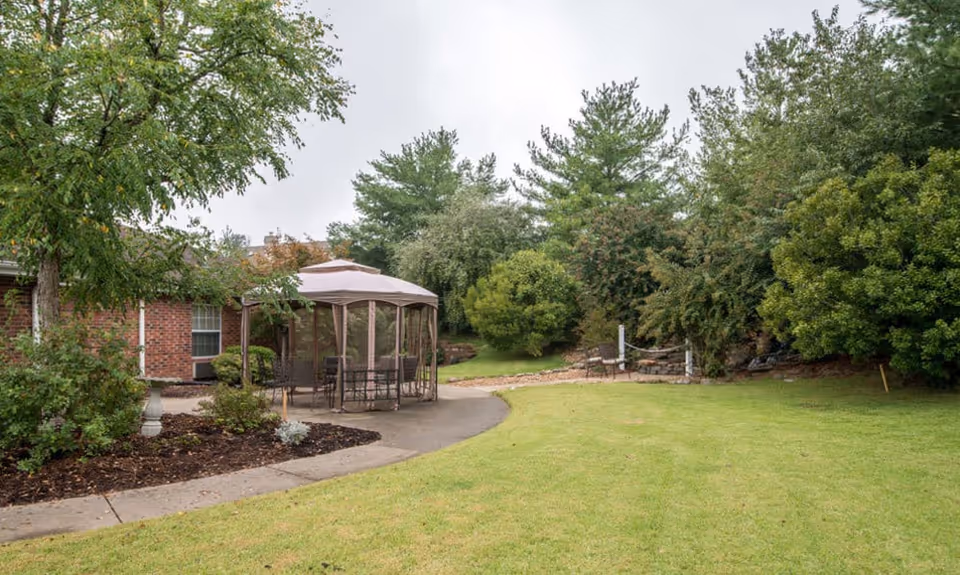 Outdoor garden area at Auburn Creek Senior Living featuring a grassy lawn, a paved walkway, a gazebo with outdoor seating, and surrounded by various trees and shrubs.