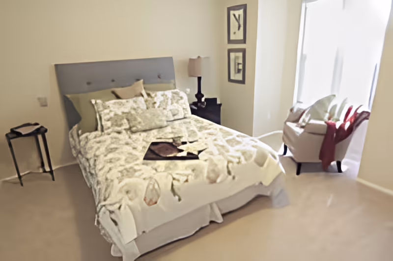 A bedroom with a neatly made bed featuring a floral patterned comforter and multiple pillows. There is a small black side table with a lamp on the right side of the bed and a small round table on the left side. A beige armchair with a red throw blanket is positioned near a window with white curtains, allowing natural light into the room. The walls are light-colored, and two framed pictures hang on the wall near the lamp.