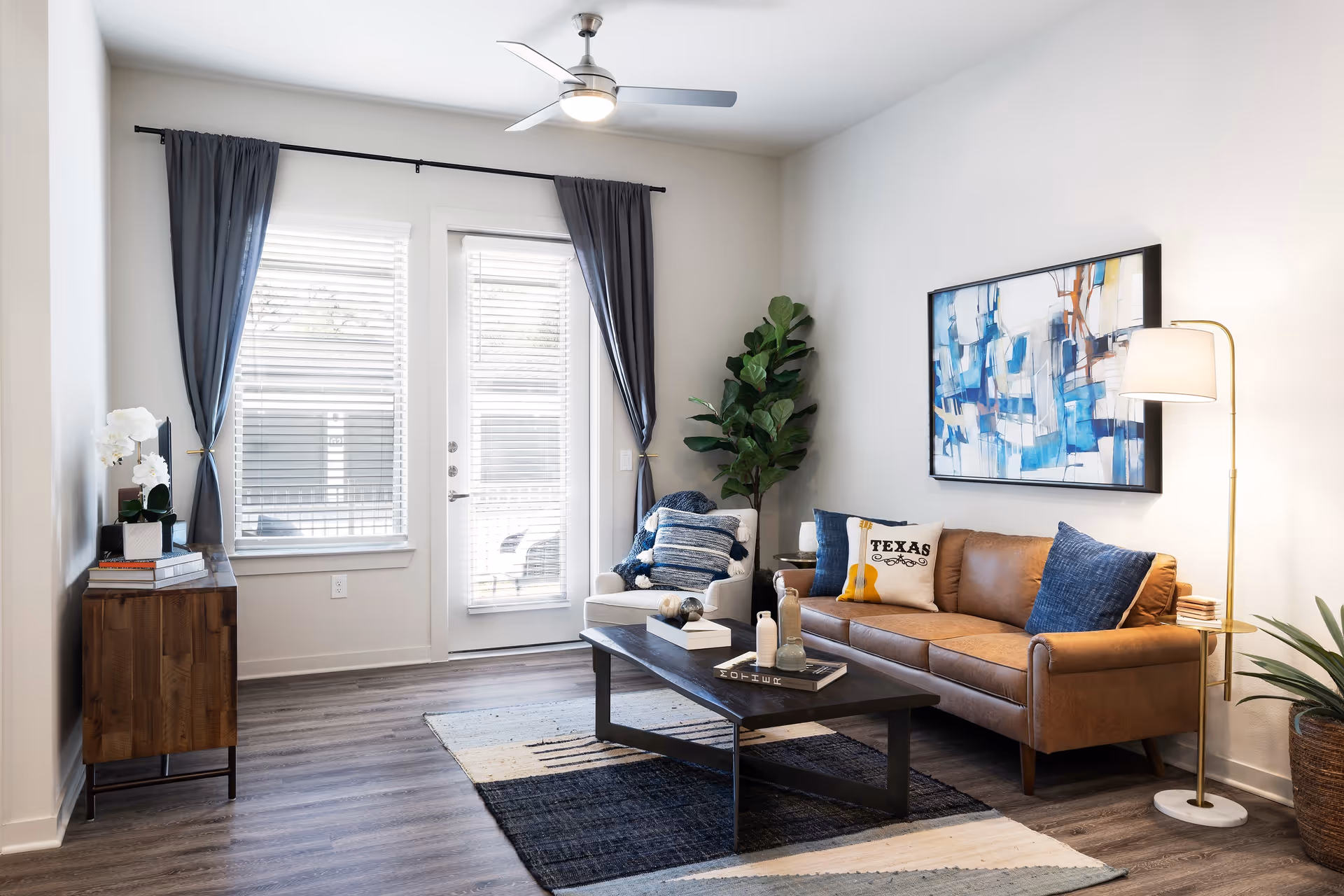 Bright modern living room with a leather sofa, armchair, coffee table, abstract wall art, and a glass door with curtains.