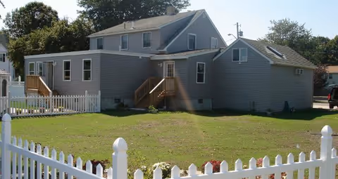 Gray multi-section two-story house with a white picket fence and a large grassy yard.