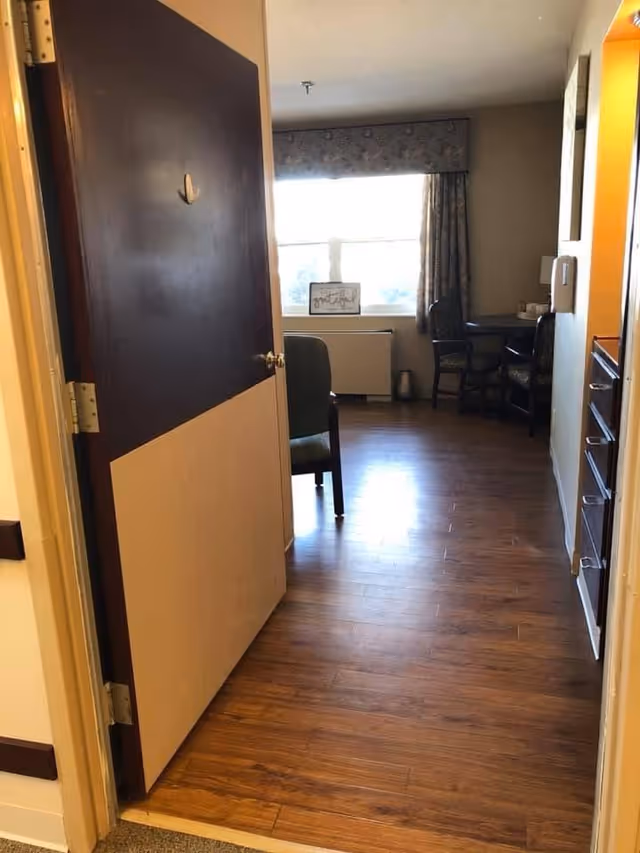 Open doorway into a senior living facility room with wood floors, chairs, a small table and a bright window with curtains.