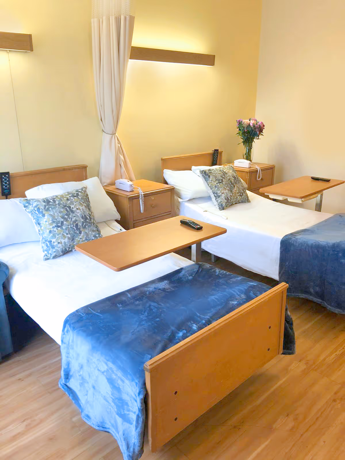 A bright room with two single beds, each with white sheets, floral pillows, and blue blankets. Each bed has a wooden overbed table with a remote control on it. Between the beds are two wooden nightstands, one with a white telephone and the other with a vase of flowers. The room has light-colored walls and wooden flooring.