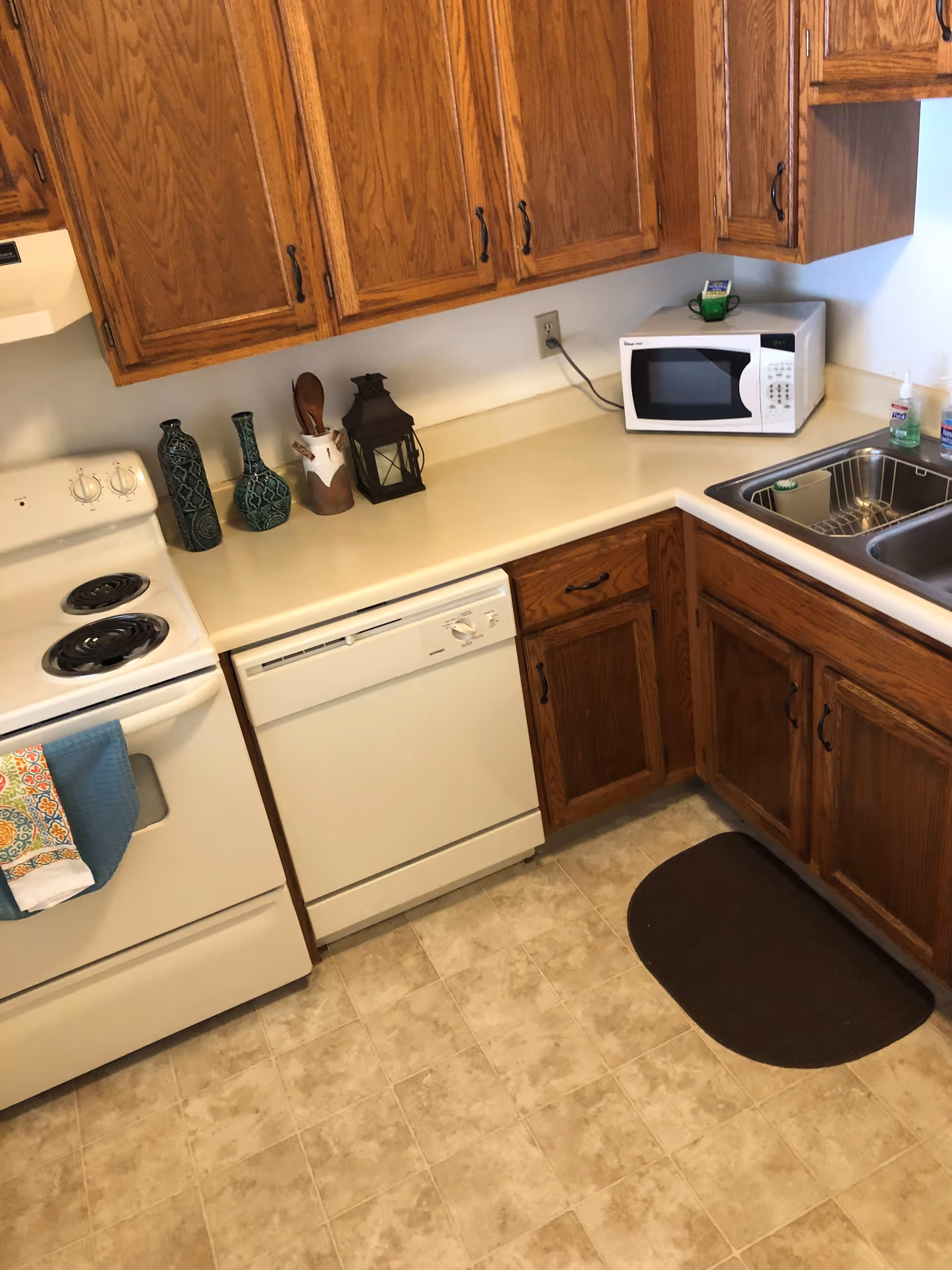 A kitchen corner with wooden cabinets, a white electric stove with two towels hanging on the handle, a white dishwasher, a white microwave on the countertop, a double stainless steel sink, and decorative items including two green vases, a utensil holder, and a lantern. The floor has beige tiles and there is a dark brown mat in front of the sink.