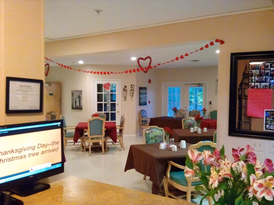 Interior view of a dining area in a senior living facility decorated with red heart garlands. Several tables are covered with brown and red tablecloths, each with chairs around them and small flower arrangements on top. A computer monitor is visible in the foreground displaying text. The room has light-colored walls and doors with windows leading outside.