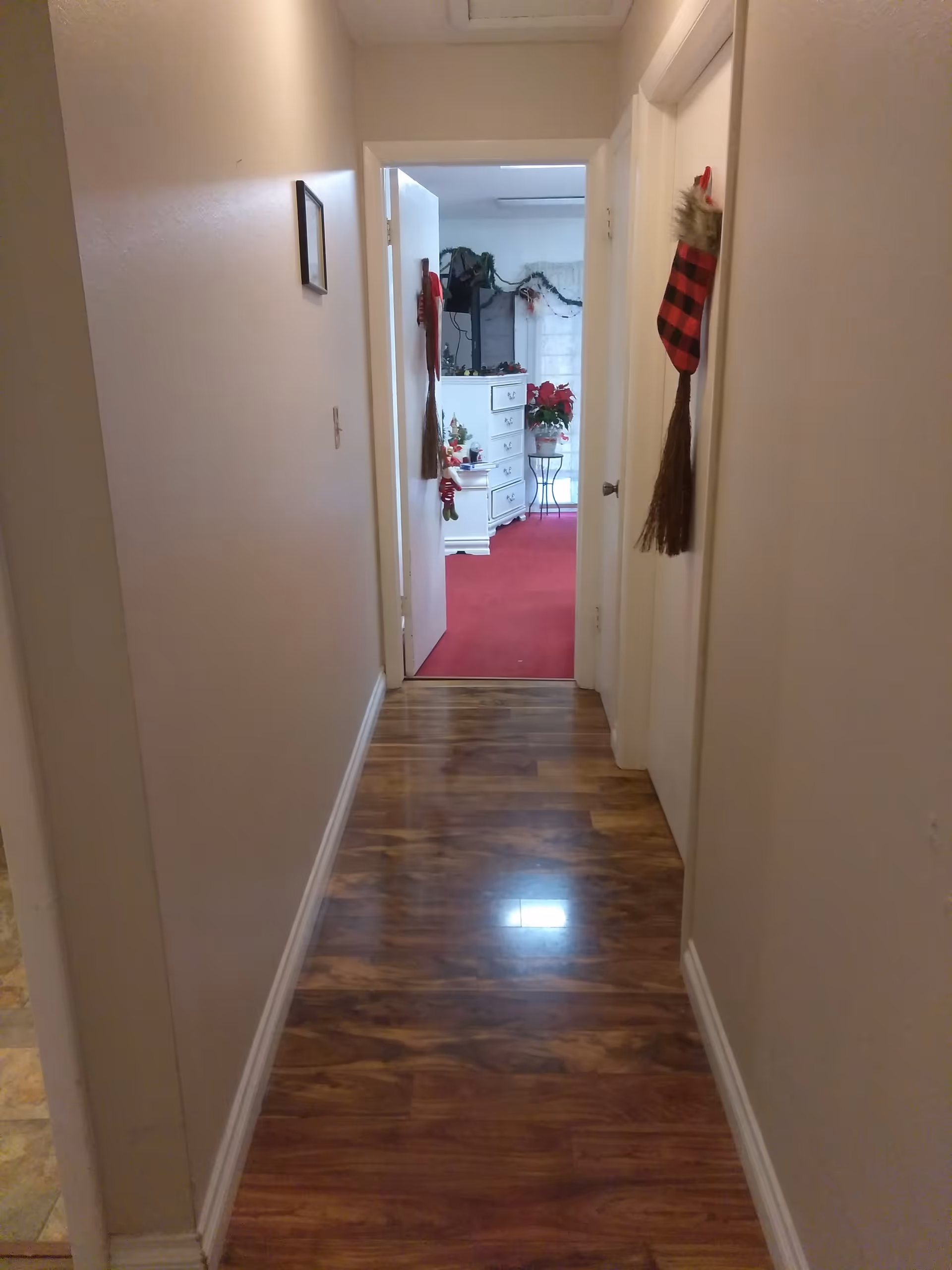 A narrow hallway with wooden flooring leading to a room with red carpet. The hallway walls are plain with a small framed picture on the left wall. Two doors are visible on the right side of the hallway, each decorated with a red and black checkered Christmas stocking with a broom attached. The room at the end of the hallway has white furniture, holiday decorations including garlands and poinsettias, and a window with sheer curtains.
