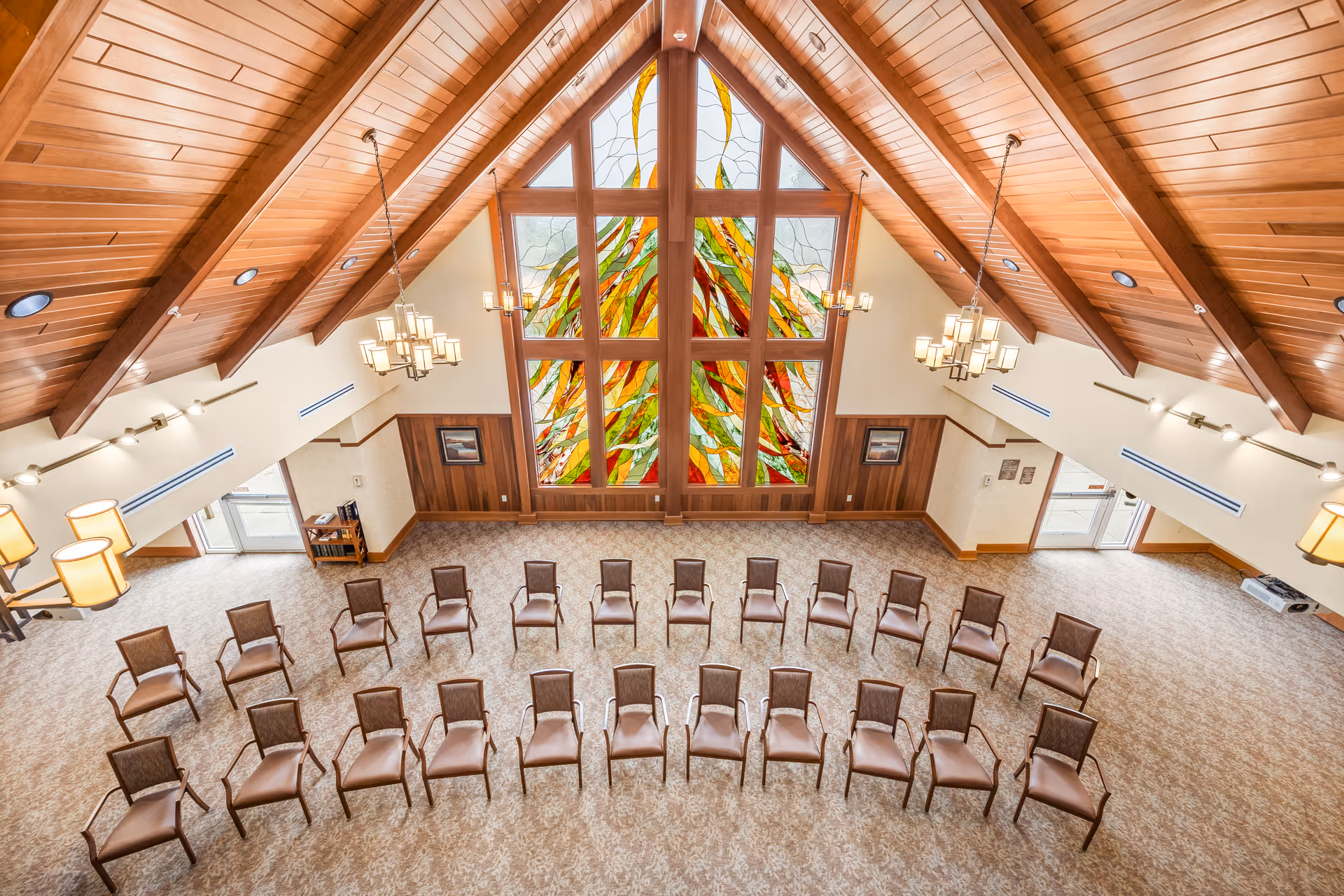 A spacious room with a high wooden vaulted ceiling and large stained glass windows featuring colorful abstract designs. The room has rows of brown chairs arranged in a semi-circle on a patterned carpet. Multiple chandeliers hang from the ceiling, and there are doors and windows on the side walls.