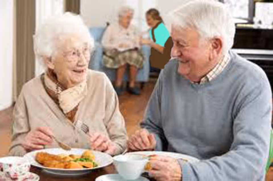 Two elderly residents smiling and eating a meal together at a dining table in a senior living facility.