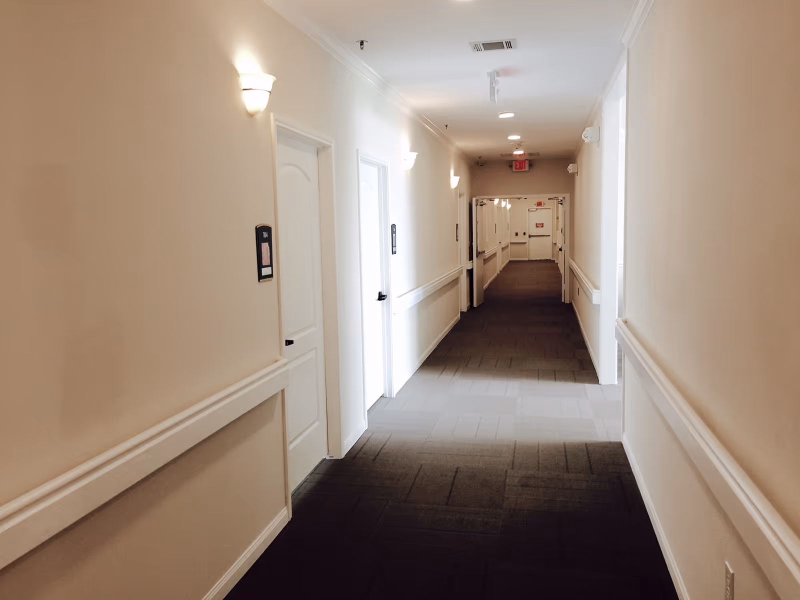 A long, well-lit hallway in a senior living facility with beige walls, white doors, wall-mounted lights, and dark carpeted flooring. The hallway has handrails on both sides and leads to a set of double doors at the end.