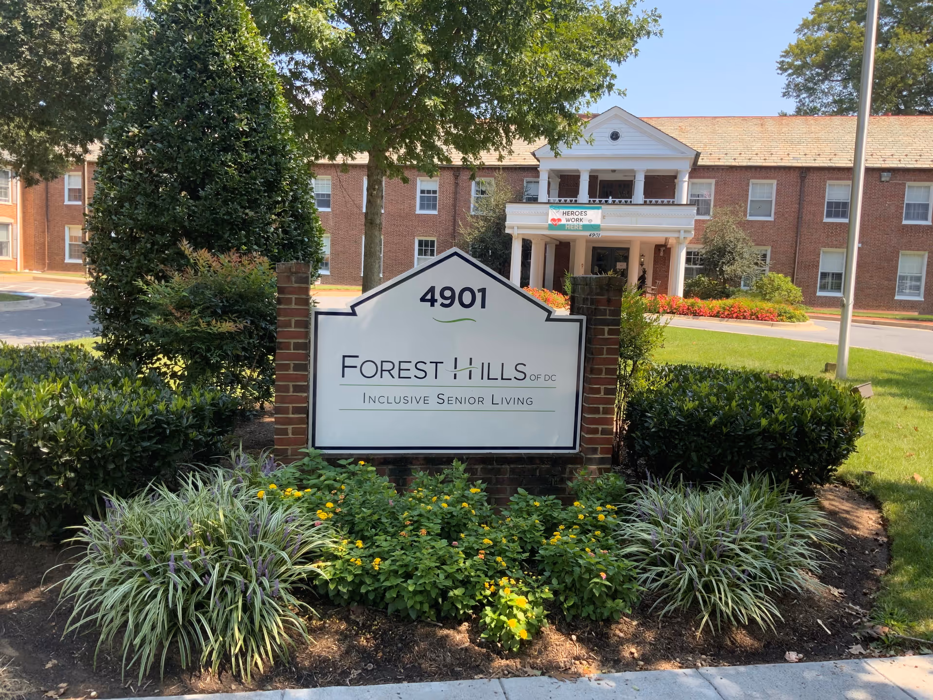 Front exterior view of Forest Hills of DC senior living facility with a large sign displaying the address 4901 and the name Forest Hills of DC Inclusive Senior Living. The building is a two-story brick structure with white columns at the entrance, surrounded by greenery and flowers.