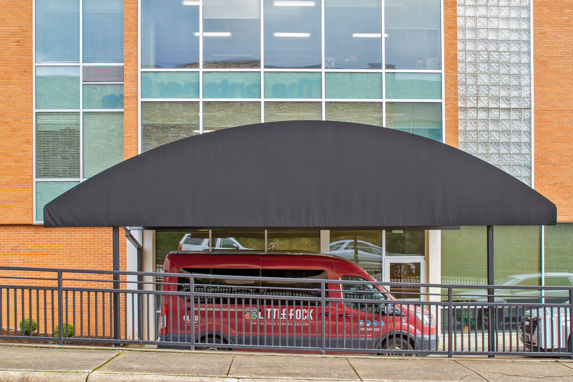 Front entrance of a brick building with a large black awning and a red shuttle van parked beneath it.